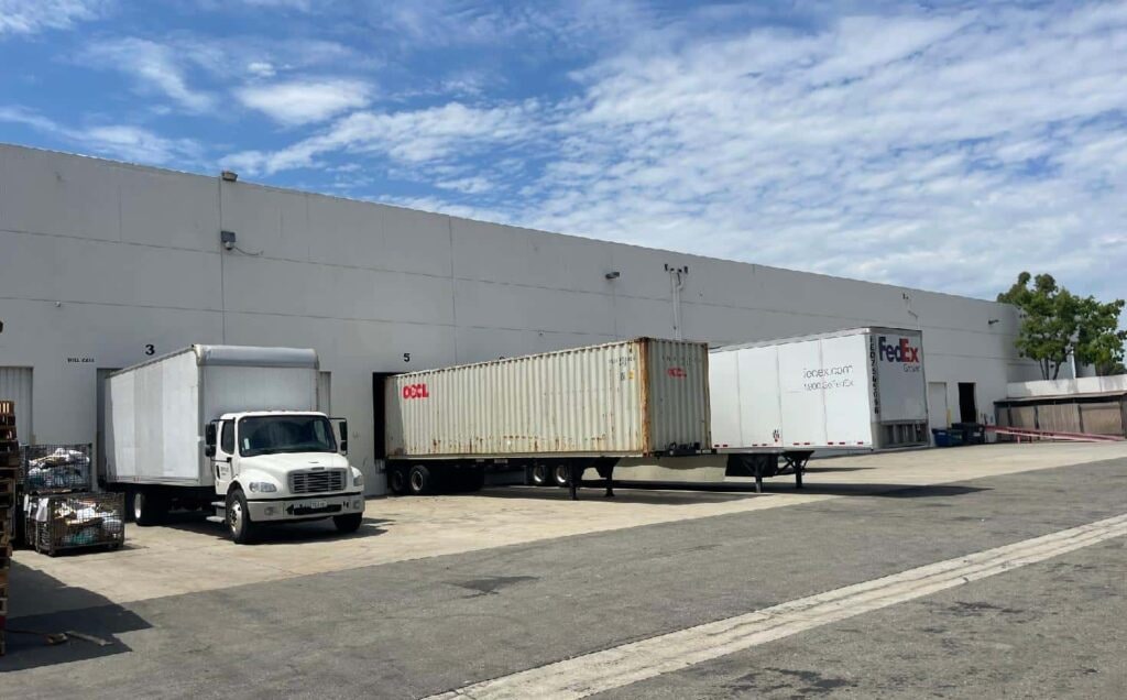 A box truck, shipping container and trailer beside warehouse loading bays